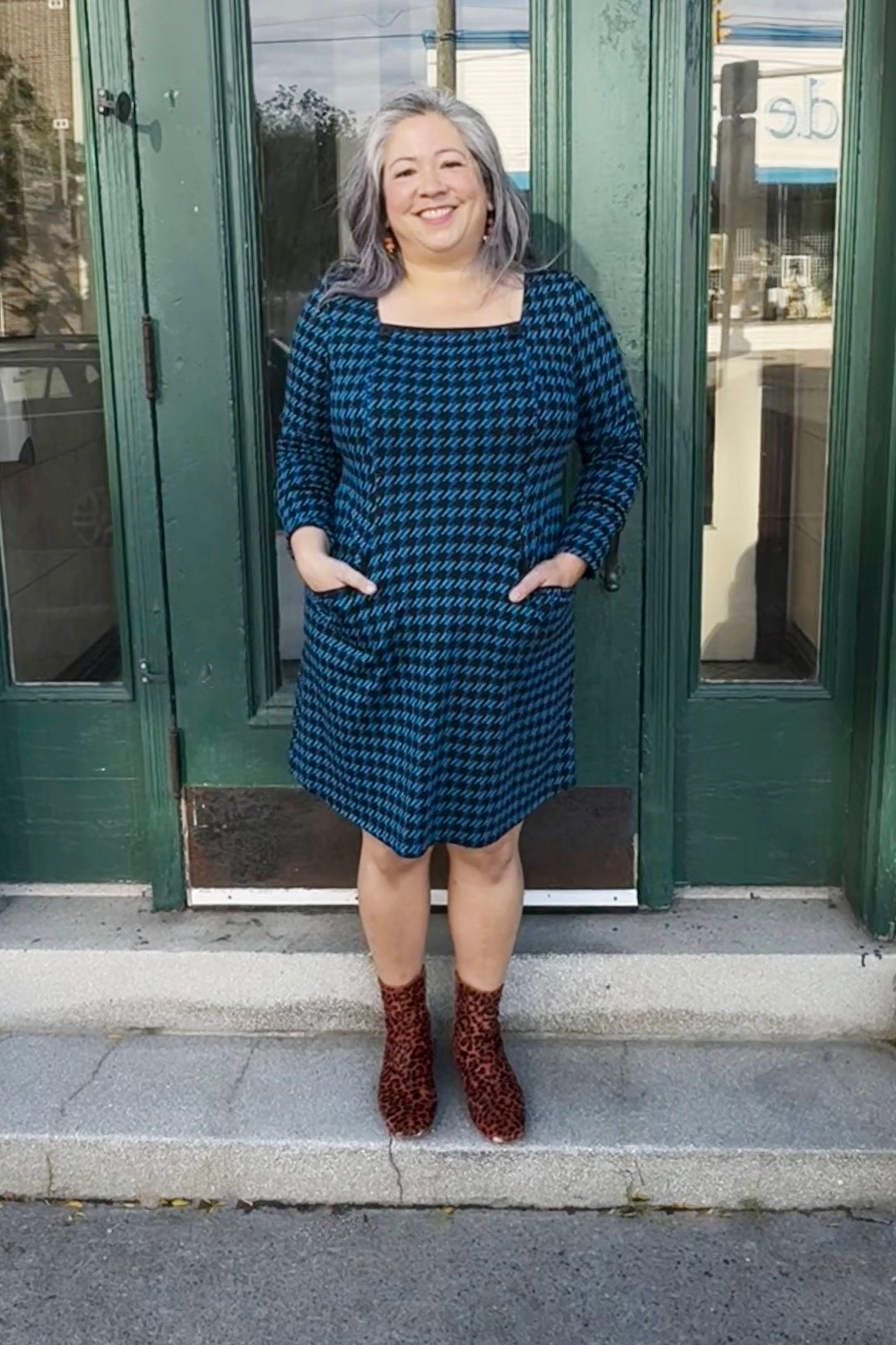 A woman wearing the Charing Dress by Luc Fontaine in Blue houndstooth, featuring a square neckline with button details, patch pockets, and a A-line shape. She is standing in front of a green doorway. 