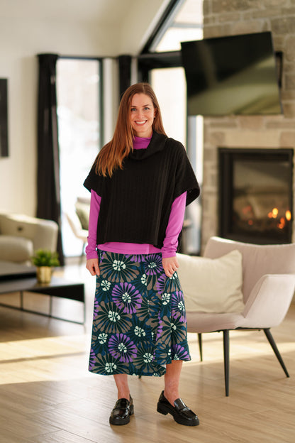 A woman wearing the Grandmont Bolero by Rien Ne Se Perd in Black,  a ribbed cowl-neck poncho. She is wearing it  over the Ste Croix Sweater in Magenta and the floral Hudson Skirt and standing in a softly lit living room with a fireplace in the background.