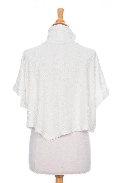 A back view of the Lupin Poncho-Bolero in Ivory by Rien ne se Perd, featuring soft jacquard knit, a loose turtleneck, and an asymmetrical hem, is shown on a mannequin in front of a white background. 