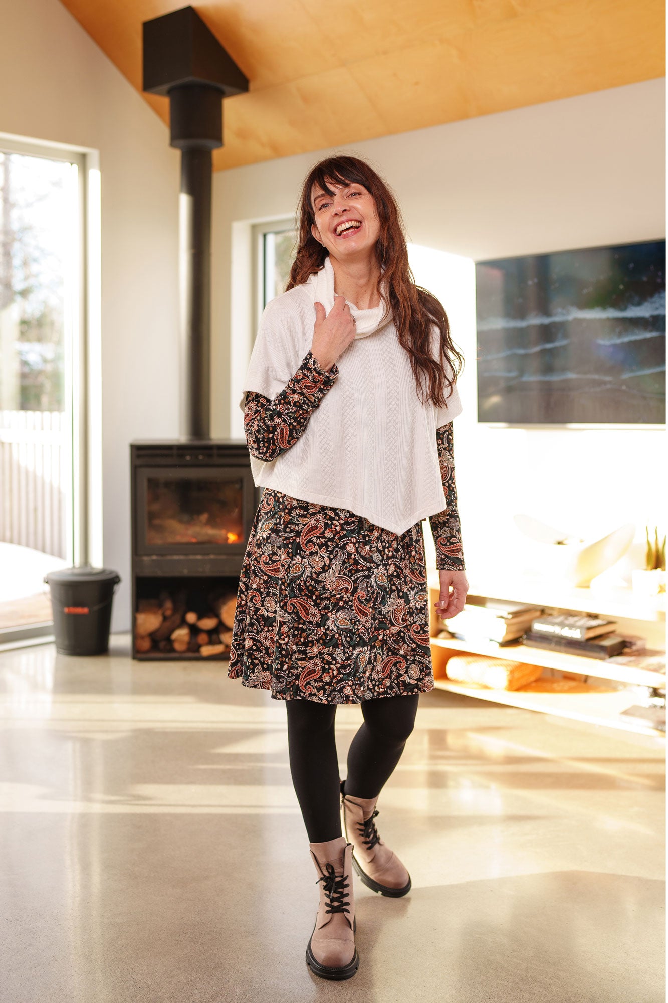 A woman wearing the Epilobe Dress by Rien ne se Perd in Fall Paisley, a classic wrap dress with a tie at the waist, delicate pleats at the shoulders, and a hem that hits above the knee. She is wearing the Lupin Poncho-Bolero in Ivory over it and is standing in a living room with a fireplace and TV.