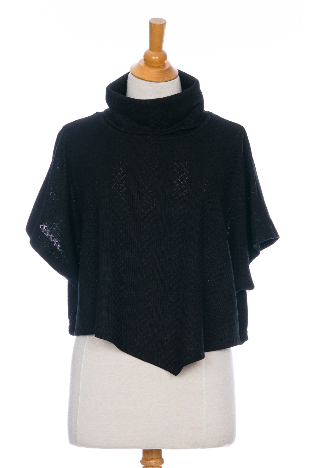 The Lupin Poncho-Bolero in Black by Rien ne se Perd, featuring soft jacquard knit, a loose turtleneck, and an asymmetrical hem, is shown on a mannequin in front of a white background. 