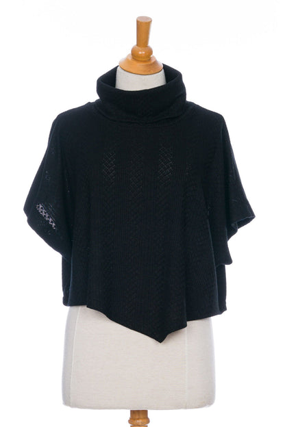 The Lupin Poncho-Bolero in Black by Rien ne se Perd, featuring soft jacquard knit, a loose turtleneck, and an asymmetrical hem, is shown on a mannequin in front of a white background. 