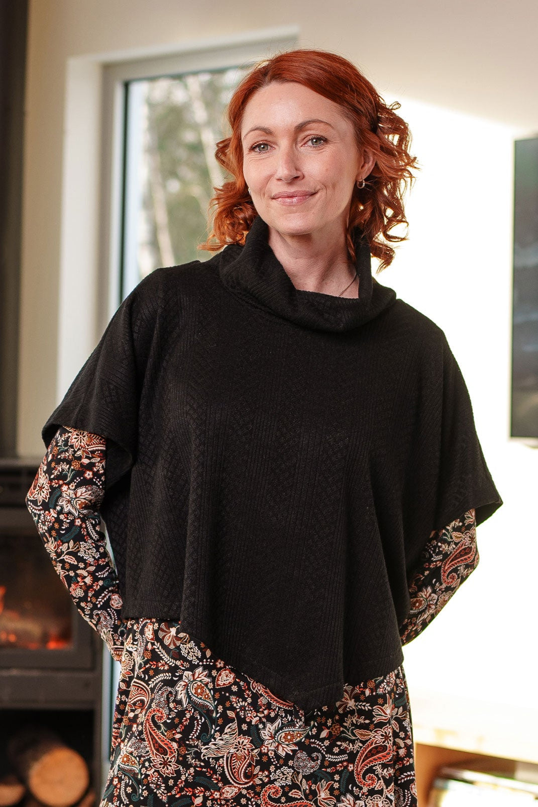 A woman wearing the Lupin Poncho-Bolero by Rien ne se Perd in Black, featuring soft jacquard knit, a loose turtleneck, and an asymmetrical hem. She is wearing it over a paisley dress and is standing in a room with a fireplace and television.

 