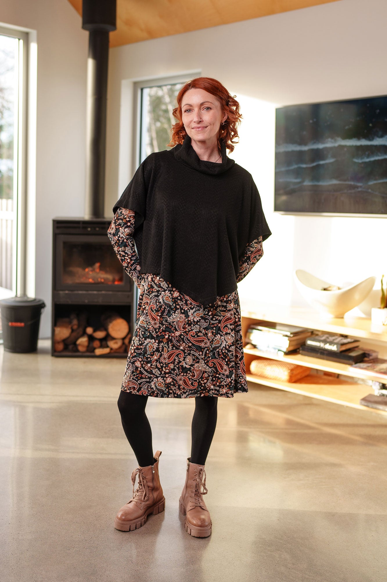 A woman wearing the Lupin Poncho-Bolero in Black by Rien ne se Perd, featuring soft jacquard knit, a loose turtleneck, and an asymmetrical hem. She is wearing it over a paisley dress and is standing in a room with a fireplace and television.