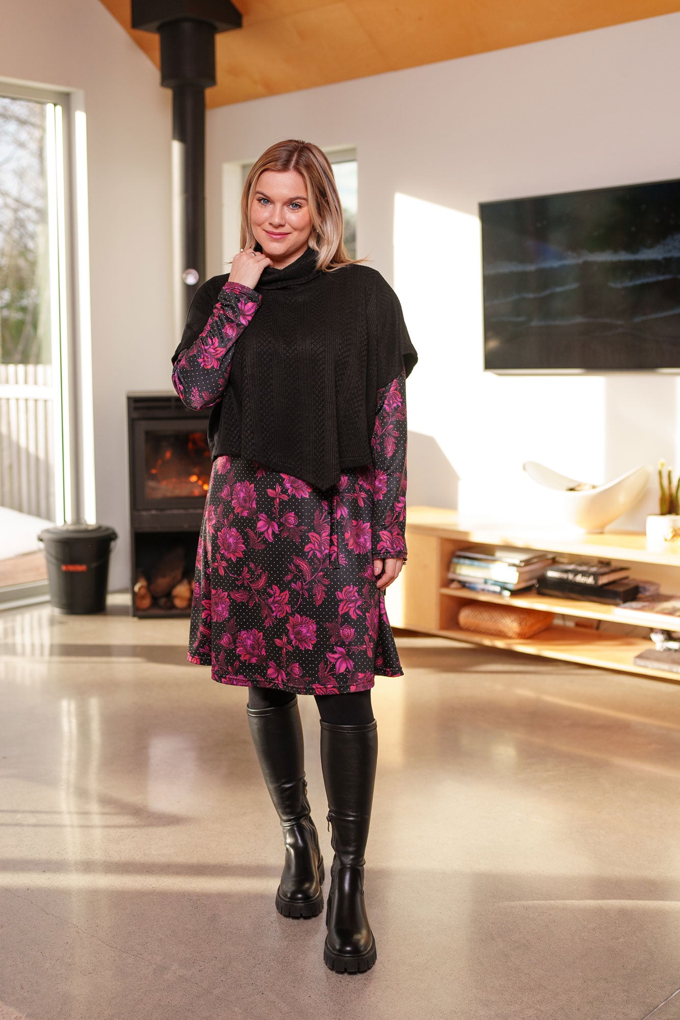 A woman wearing the Lupin Poncho-Bolero in Black by Rien ne se Perd, featuring soft jacquard knit, a loose turtleneck, and an asymmetrical hem. She is wearing it over a pink floral dress and is standing in a room with a fireplace and television.