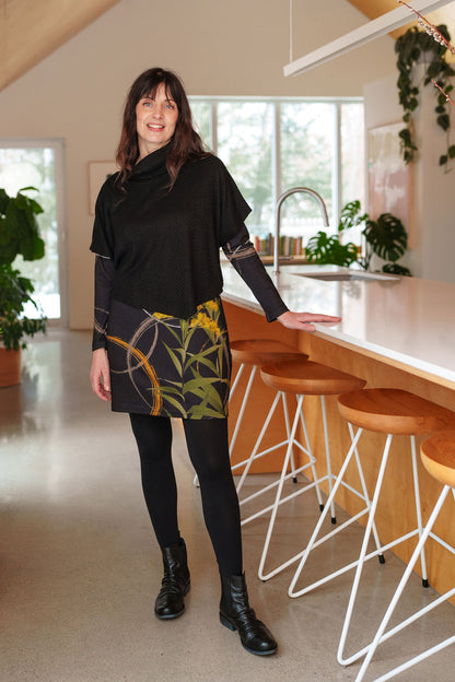 A woman wearing the Lupin Poncho-Bolero in Black by Rien ne se Perd, featuring soft jacquard knit, a loose turtleneck, and an asymmetrical hem. She is wearing it over leaf patterned tunic and is standing in a kitchen.
