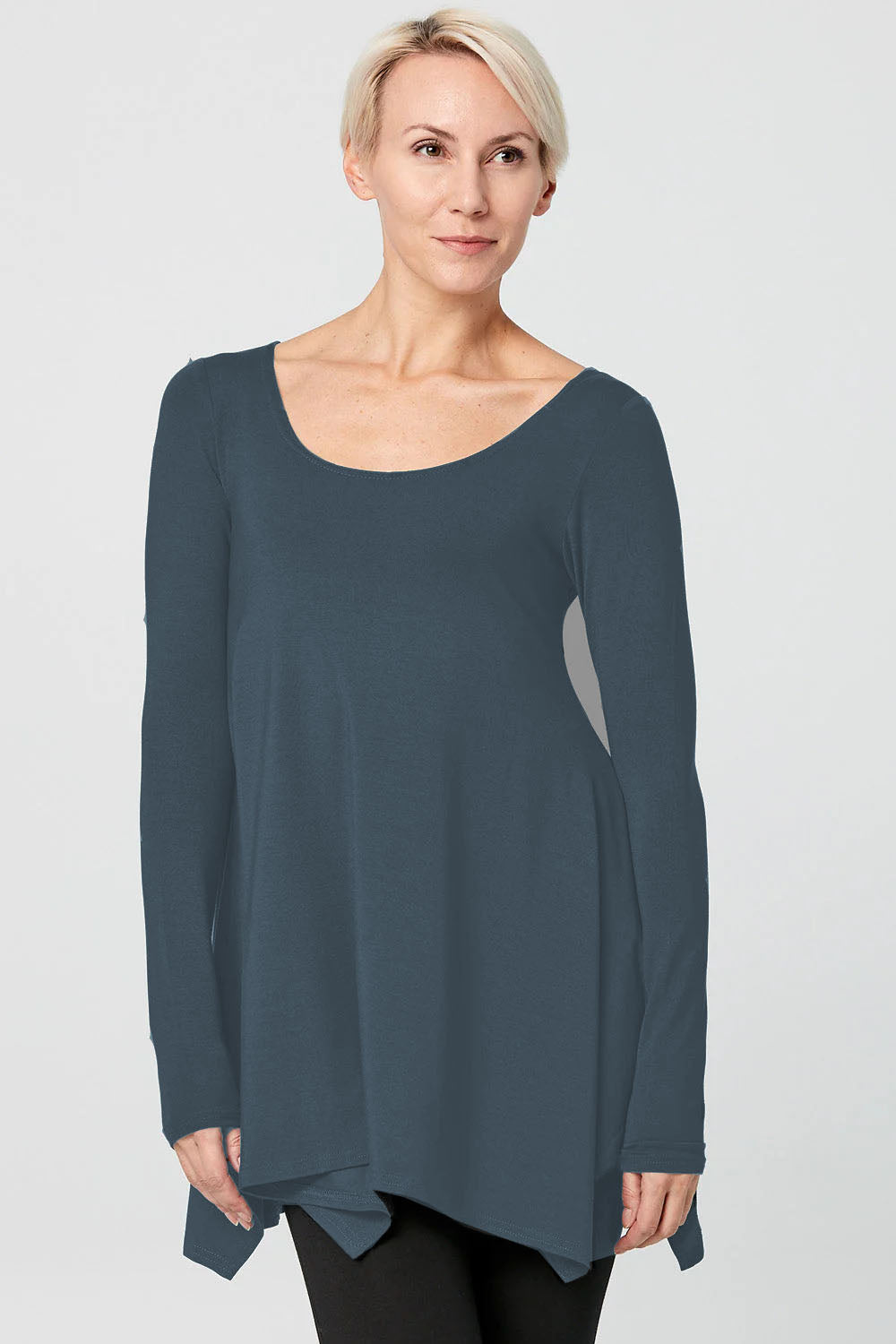 A woman wearing the Breath Top by Advika in Steel, featuring a scooped neck, long sleeves, and a swing shape with an asymmetrical hem, all in organic cotton/tencel. She is wearing it with leggings and standing in front of a white background. 