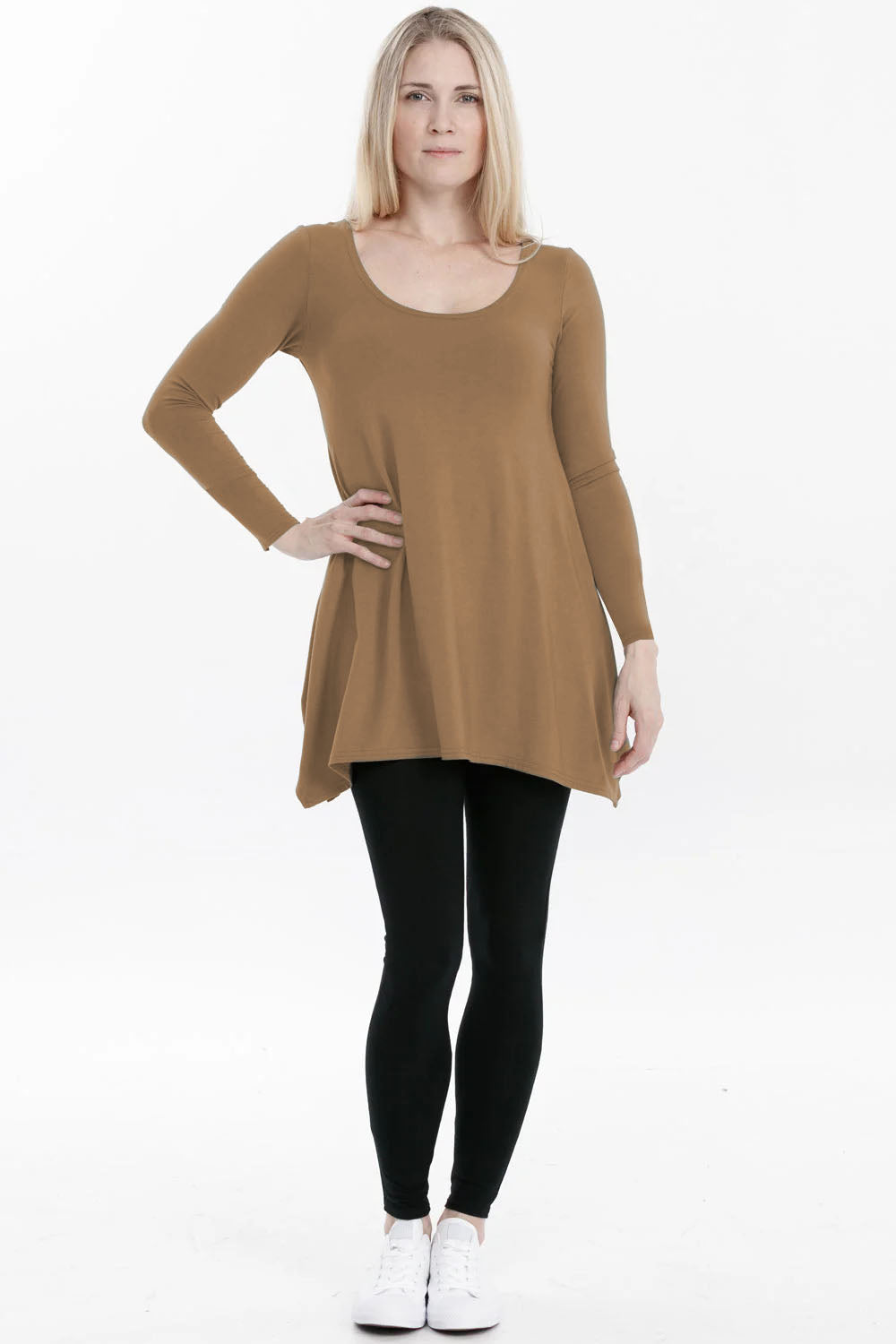 A woman wearing the Breath Top by Advika in Whiskey, featuring a scooped neck, long sleeves, and a swing shape with an asymmetrical hem, all in organic cotton/tencel. She is wearing it with leggings and standing in front of a white background. 