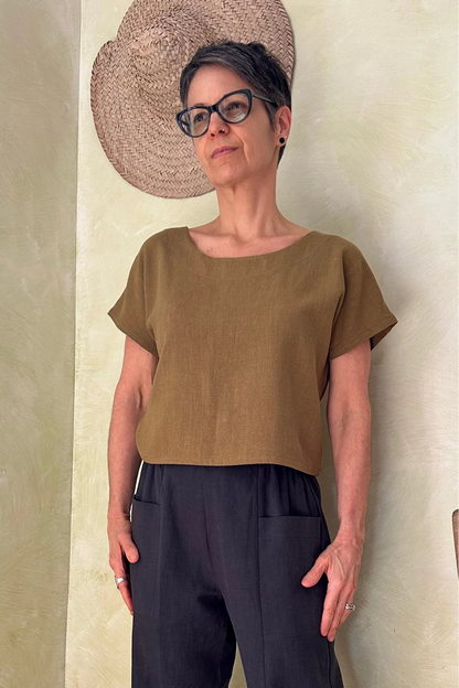 A woman wearing the Brendola Top from Kazak in Mousse, a slightly cropped short sleeved top with three brass snaps that can be worn at the back or the front. She is wearing it with the navy pants and standing in a room with hats on the wall. 