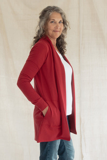 Side view of woman wearing the Briton Cardigan from Blondie in Poppy, a mid-thigh length open cardigan with a small collar and in-seam pockets. She is wearing it with a white tee shirt and jeans and standing in front of a beige background 