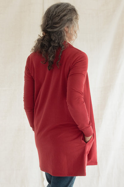 Back view of a woman wearing the Briton Cardigan from Blondie in Poppy, a mid-thigh length open cardigan with a small collar and in-seam pockets. She is wearing it with a white tee shirt and jeans and standing in front of a beige background 