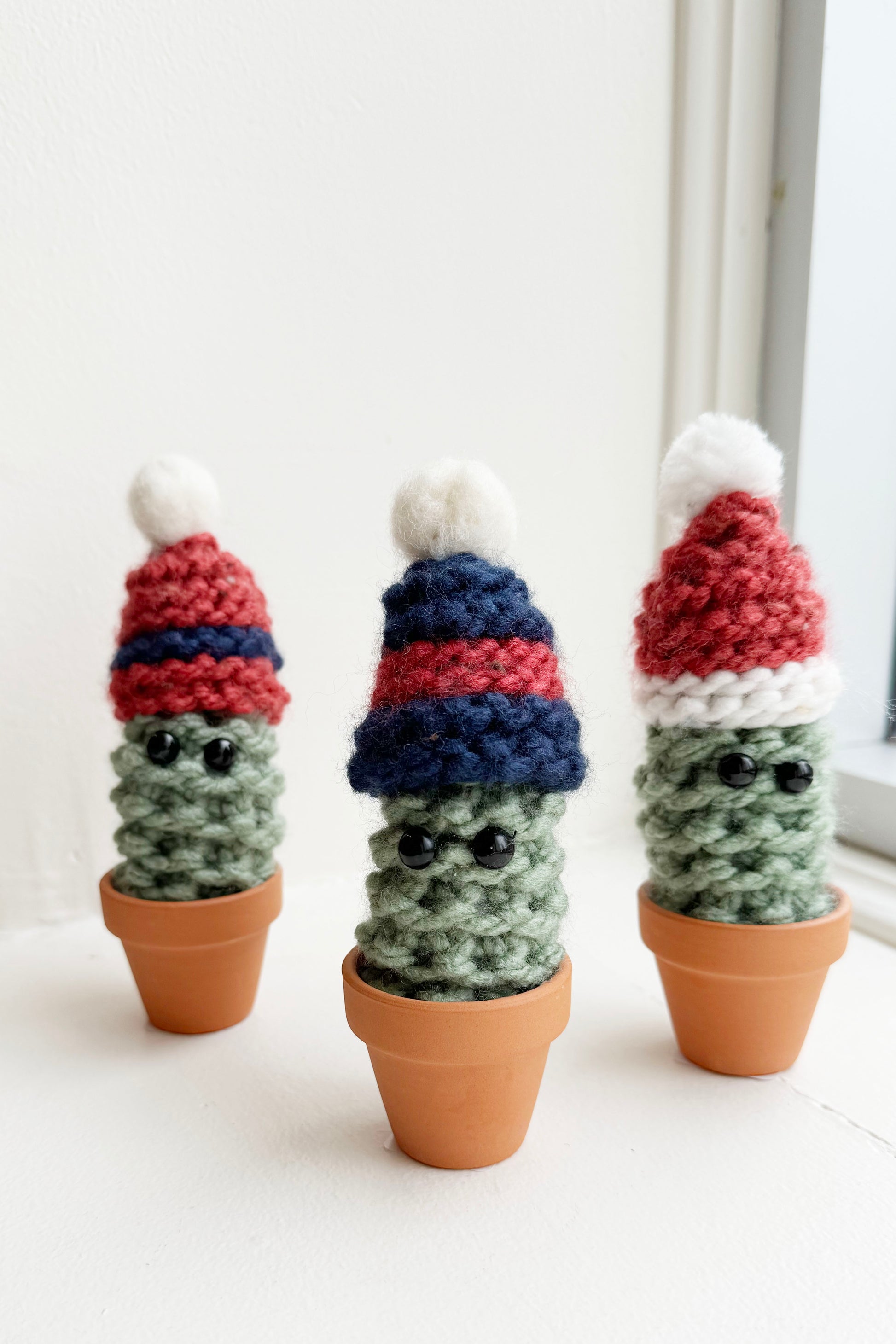 Knitted Winter Cacti by Curl Country, with hats and set in terracotta pots, are shown in front of a white background. 