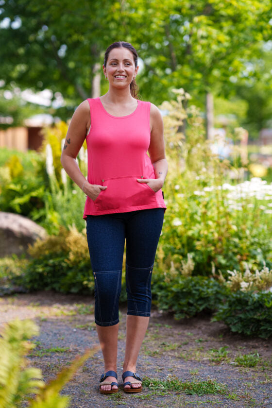 A woman wearing the LIndy Capri pants from Rien ne se Perd, faux denim stretch capris with a wide pull-on waist, contrast seams at the bottom, and decorative coconut buttons. She is wearing them with a pink tank top and standing in a garden. 