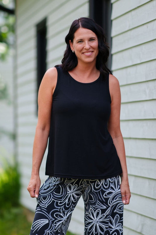 A woman wearing the Spritz Tank Top by Rien ne se Perd in Black, with wide straps and side slits. She is wearing it with  a black tank top and patterned pants and standing in front of a house.