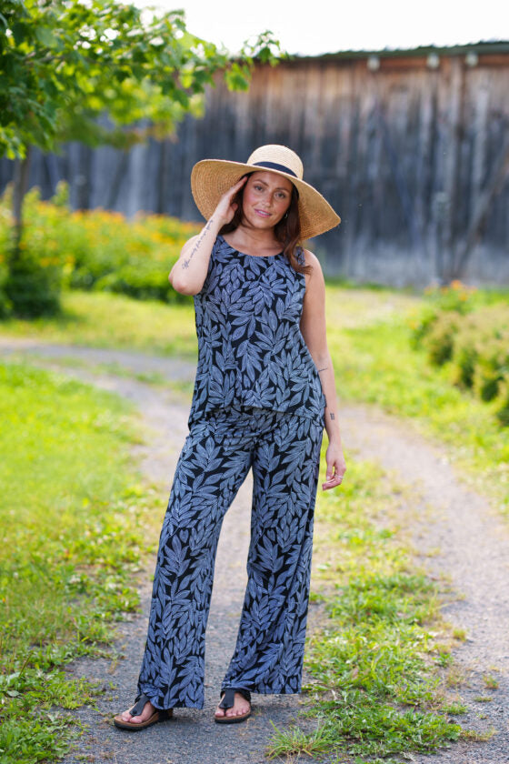 A woman wearing the Swing Cami by Rien ne se Perd in Black and White Foliage, a trapeze cut tank with a hi-low rounded hemline. She is wearing it with the matching Rumba Palazzo pants and standing outside in front of a wooden building. 