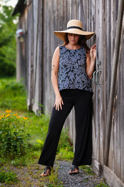 A woman wearing the Swing Cami by Rien ne se Perd in Black and White Foliage, a trapeze cut tank with a hi-low rounded hemline. She is wearing it with black pants  and standing outside in front of a wooden building. 