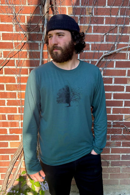 A man wearing the Camouflage T-Shirt in Teal from Message Factory, a long-sleeve tee with black tree design. He is wearing it with black pants and standing in front of a brick wall.