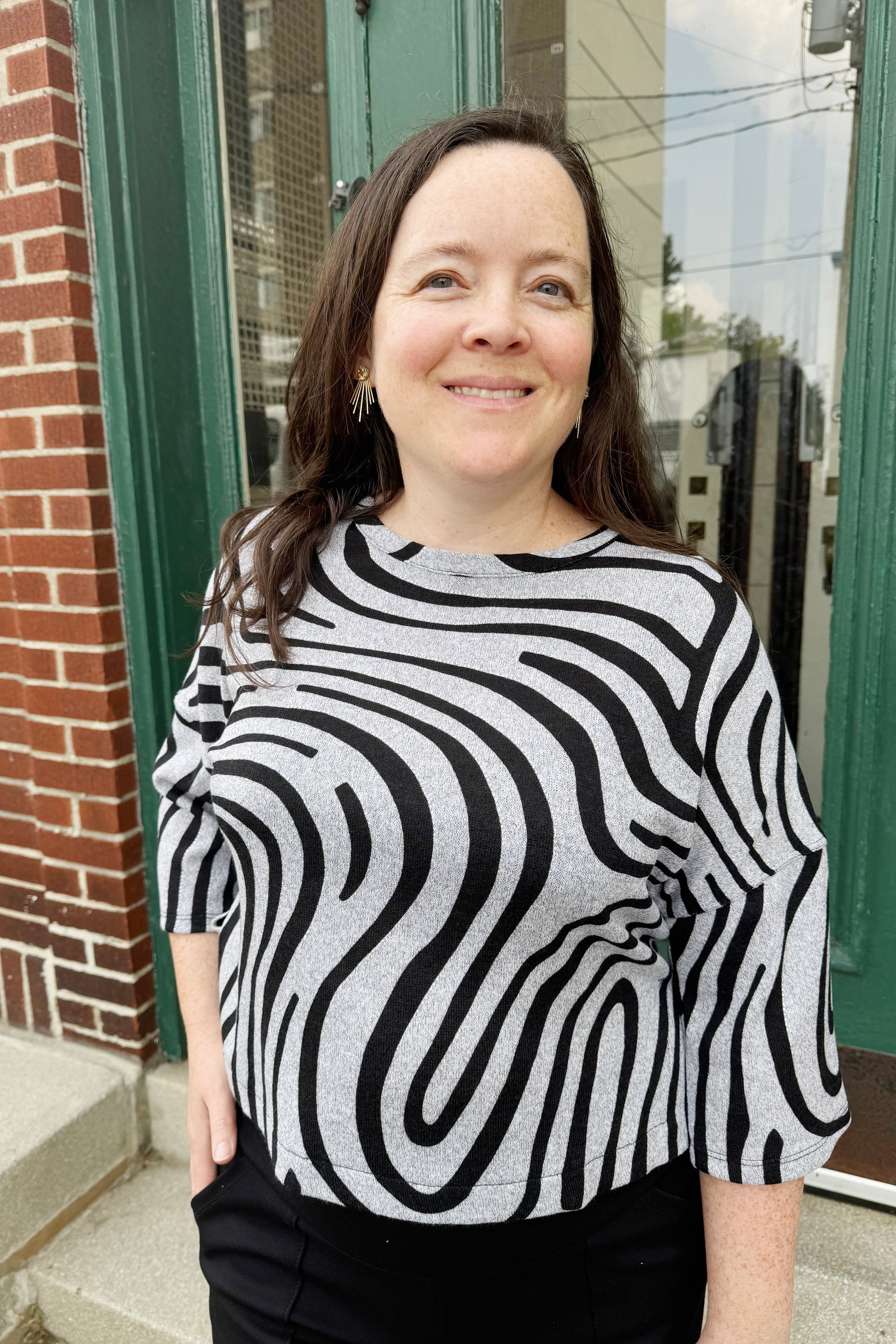 A woman wearing the Paula Top by Compli K in Silver Black Swirl, with round neck, 3/4 drop sleeves, and a hip length, with black shorts. She is standing outside in front of a door. 