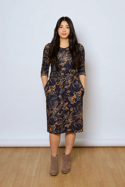 A woman wearing the Cascade Dress by Tangente in Navy Print, with a round neck, 3/4 sleeves, gathered defining waistband, and a gathered knee-length skirt. She is standing against a white wall.