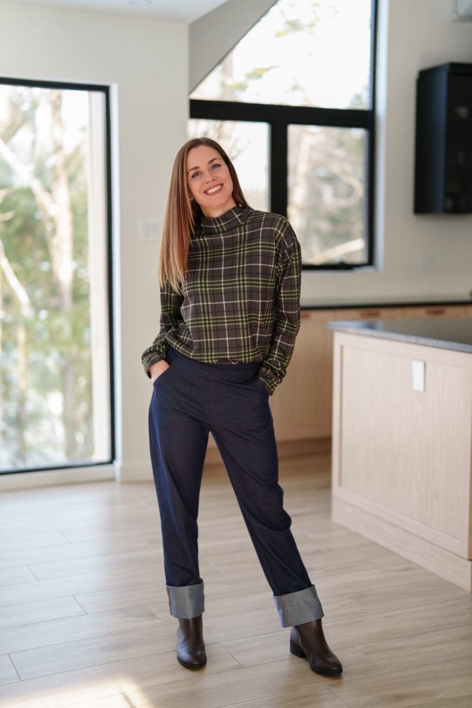 A woman wearing the Yamaska Pants by Rien ne se Perd in Denim Blue, pull on style jeans with rolled cuffs. She is wearing them with a green and brown plaid Batiscan Sweater and dark brown ankle boots in a bright room with light wood flooring