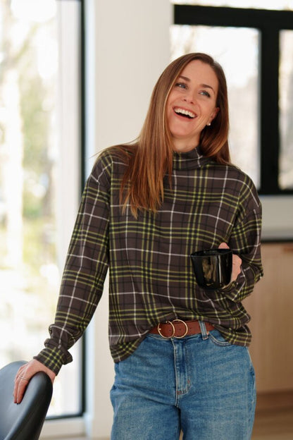 A woman wearing the green and brown plaid Batiscan Sweater by Rien ne se Perd, with a mock-neck and long sleeves. She is wearing it with jeans and standing indoors holding a  coffee mug. 