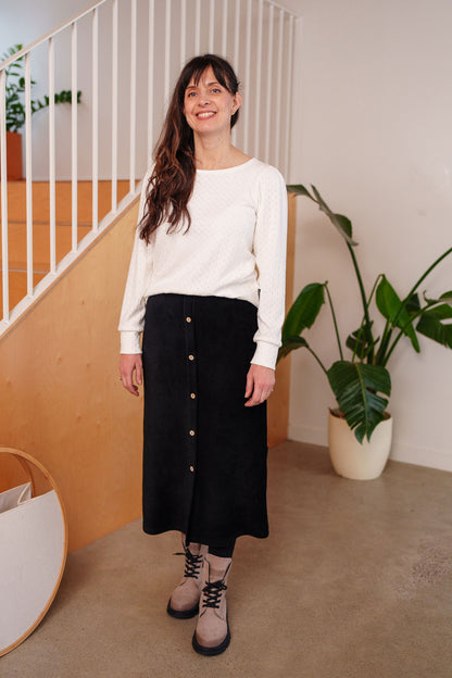 A woman wearing the Pruche Skirt by Rien ne se Perd, a 7/8 length Black corduroy skirt with an elastic waist, five coconut buttons up the front, and a slit at the back. She is wearing it with a white top and standing indoors. 