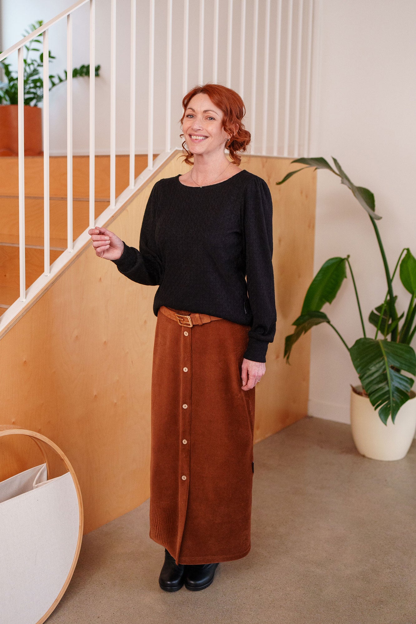 A woman wearing the Camarine Sweater by Rien ne se Perd in Black, a cable knit sweater with a scooped neck, puffed shoulders, and gathered cuffs, with a copper skirt She is standing indoors by a staircase.