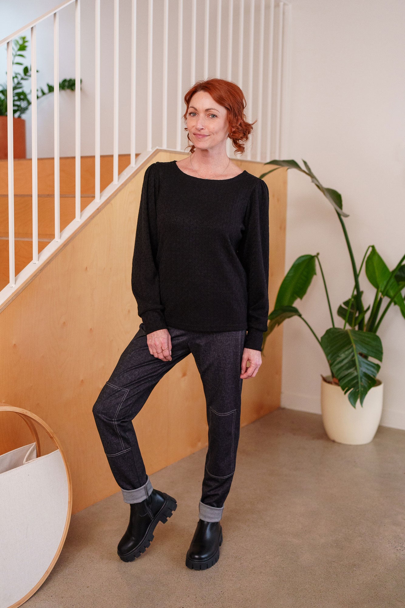 A woman wearing the Cypres Pants by Rien ne se Perd in Blue Denim, stretchy faux denim pants with a pull on waist and tone-on-tone patches on the knees. She is wearing with them with a black sweater and standing in front of a railing. 