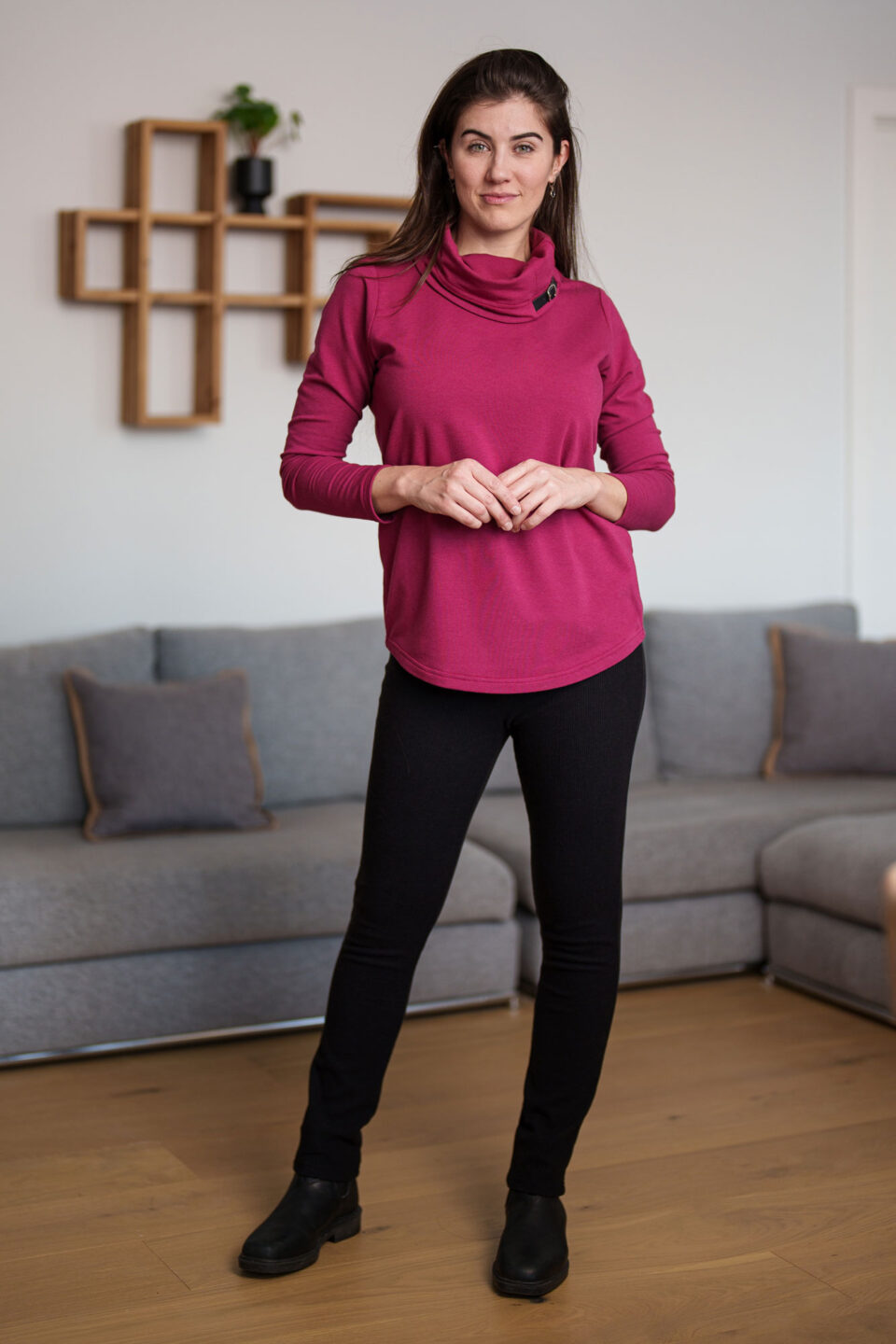 A woman wearing the Lichen Pant by Rien ne se Perd in Black, skinny leg pull-on pants in stretchy corduroy fabric. She is wearing them with a pink long-sleeve top and standing in a living room. 