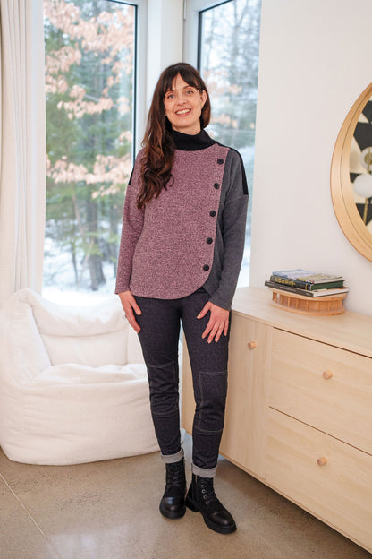 A woman wearing the Cypres Pants by Rien ne se Perd in Blue Denim, stretchy faux denim pants with a pull on waist and tone-on-tone patches on the knees. She is wearing them with a sweater and standing in front of a white sofa. 