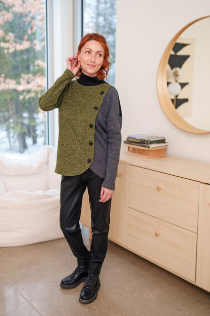 A woman wearing the Chèvrefeuille Sweater by Rien ne se Perd in Sage, a multi-toned piece that's green on the right side, grey on the left, with a black back. It has a cross-over front with five black buttons. She is wearing it with dark pants and standing in front of a window. 