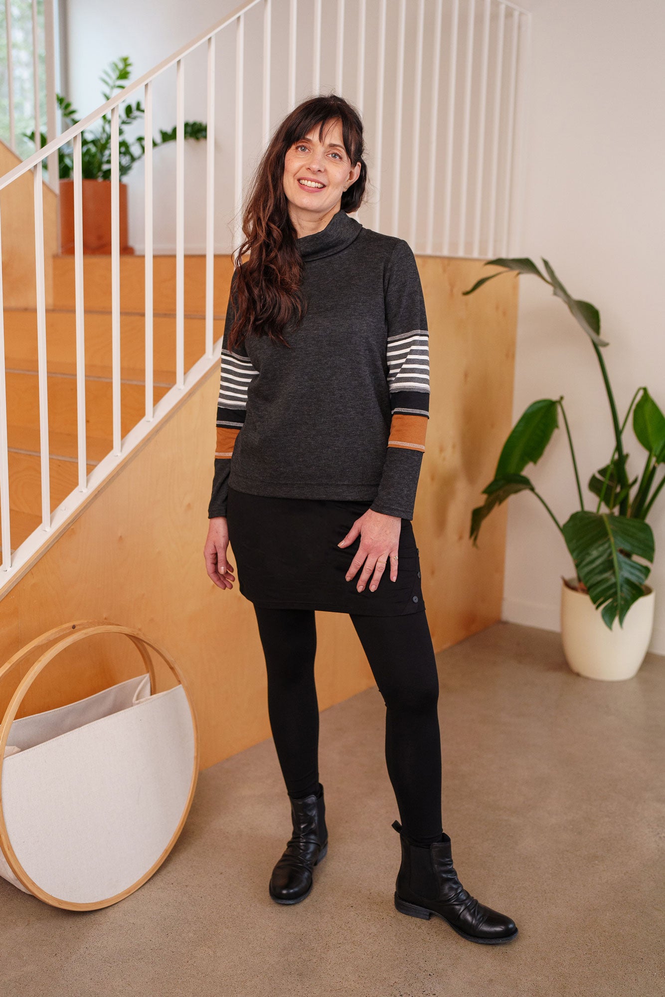 A woman wearing the Chicouté Sweater by Rien ne se Perd in Charcoal/Cognac, with a small turtleneck colour-blocked sleeves. She is wearing it with a black skirt and standing in front of a railing. 