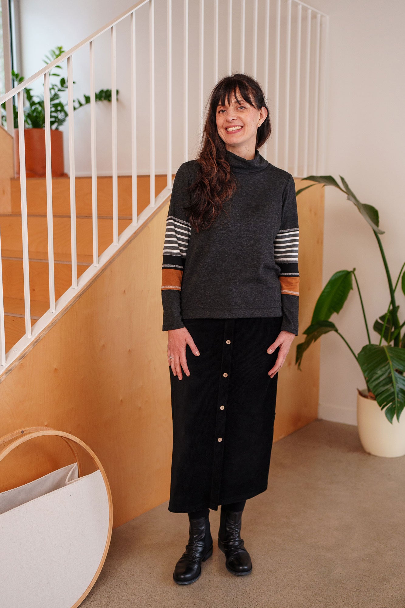 A woman wearing the Chicouté Sweater by Rien ne se Perd in Charcoal/Cognac, with a small turtleneck colour-blocked sleeves. She is wearing it with the Pruche skirt in Black and standing in front of a railing. 