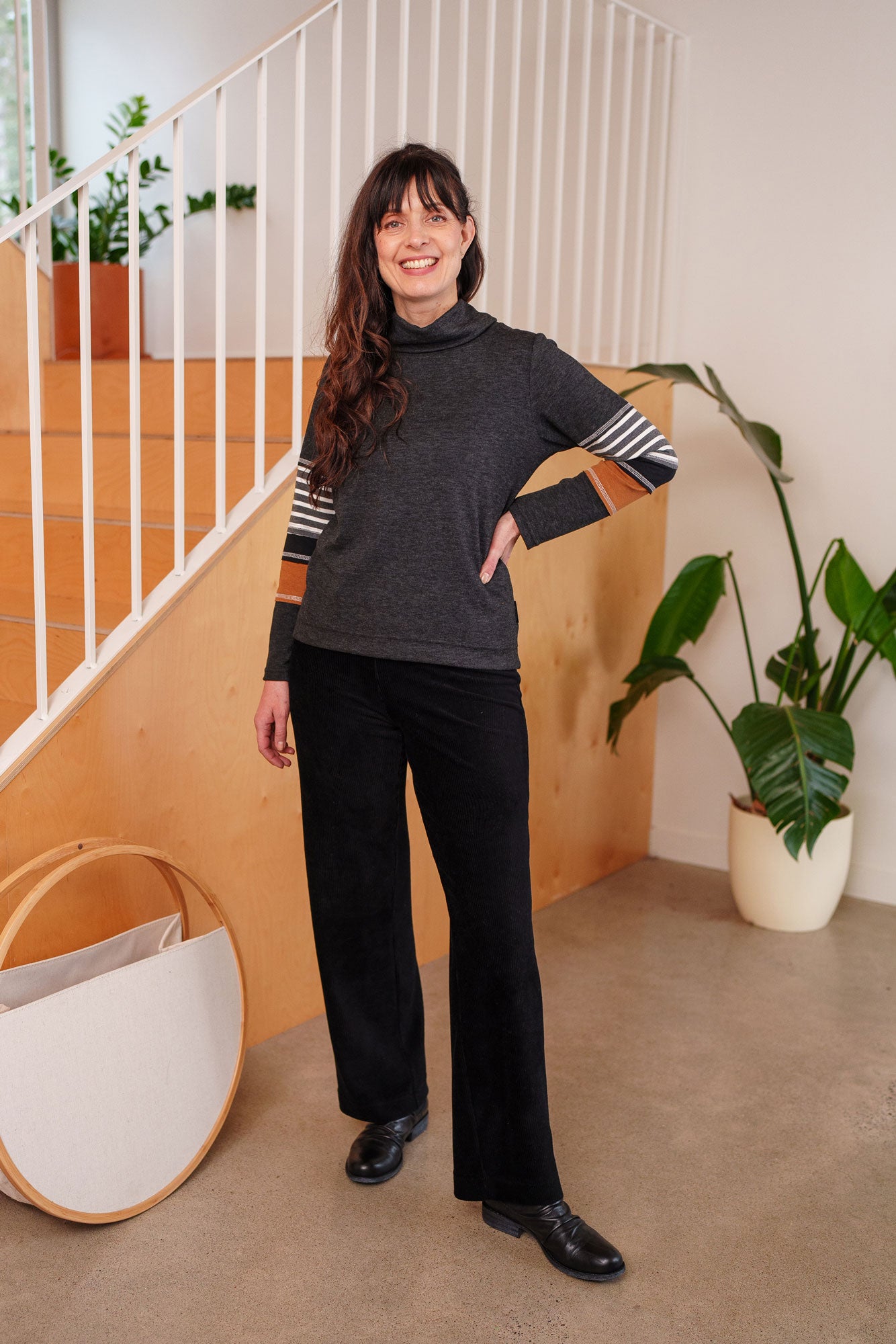 A woman wearing the Chicouté Sweater by Rien ne se Perd in Charcoal/Cognac, with a small turtleneck colour-blocked sleeves. She is wearing it with black pants and standing in front of a railing. 
