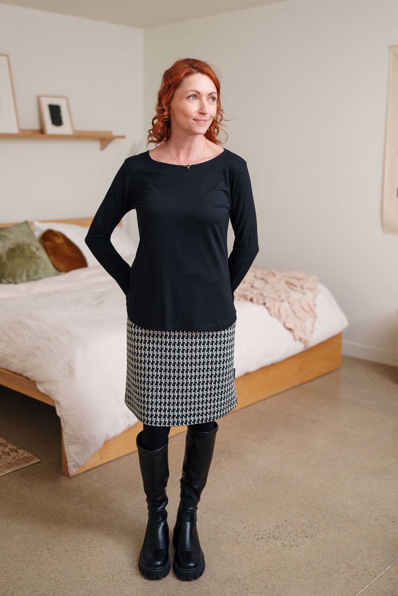 A woman wearing the Meleze Skirt by Rien ne se Perd in Black and White houndstooth,  a knee-length straight skirt with an elastic waist, with a black top. She is standing in a bedroom.