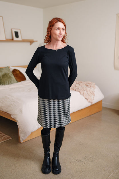 A woman wearing the Meleze Skirt by Rien ne se Perd in Black and White houndstooth,  a knee-length straight skirt with an elastic waist, with a black top. She is standing in a bedroom.