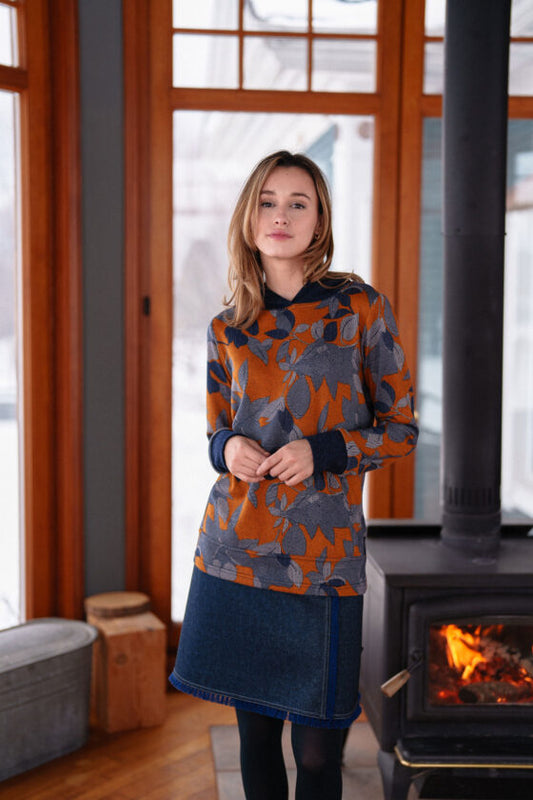 A woman wearing the Lautrec Sweater by Rien ne se Perd in Navy Foliage with a denim skirt stands in front of a wood stove. 