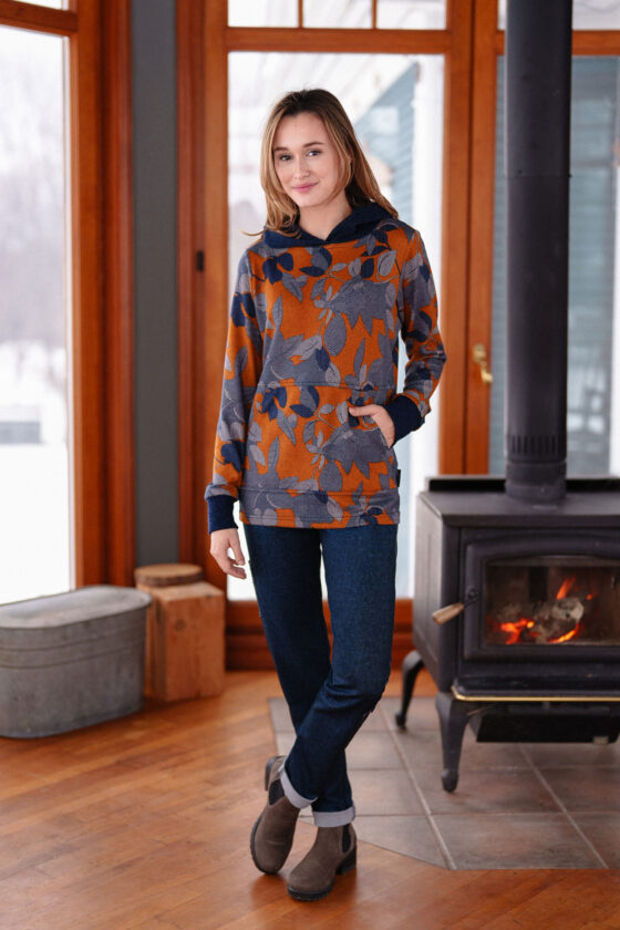 A woman wearing the Lautrec Sweater by Rien ne se Perd in Navy Foliage with jeans stands in front of a wood stove. 