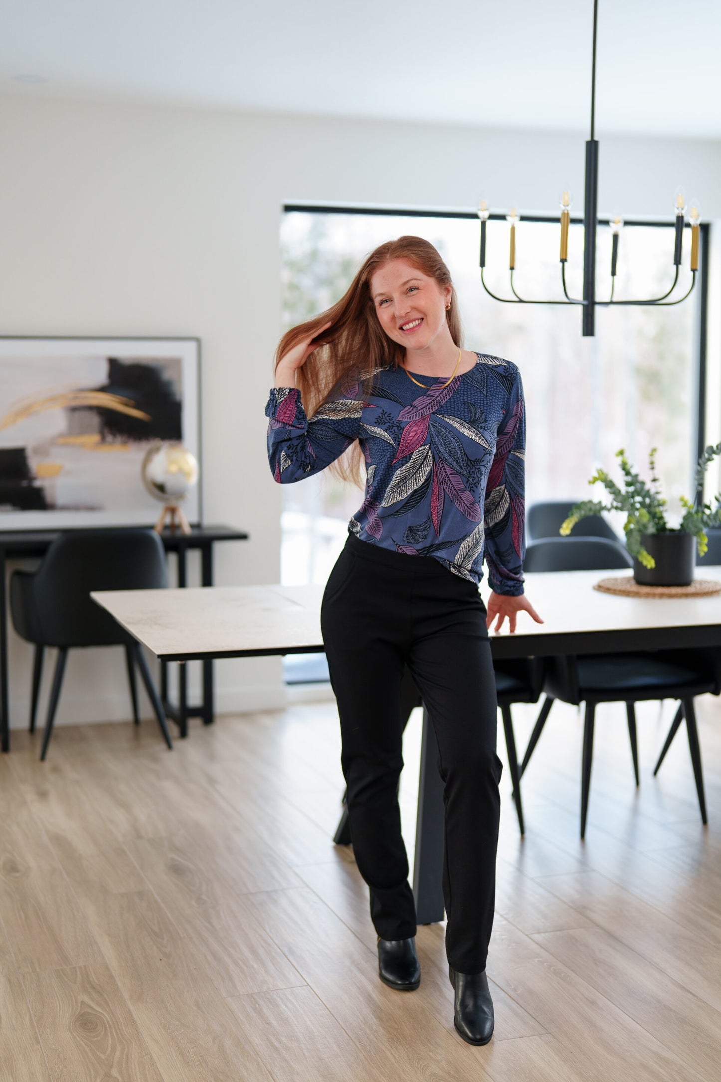 A woman wearing the St Charles Sweater by Rien ne se Perd in Foliage, a jewel toned patterned top with long sleeves and elastic cuffs. She is wearing it with the Murphy Pants in Black and 
standing in a modern kitchen. 
