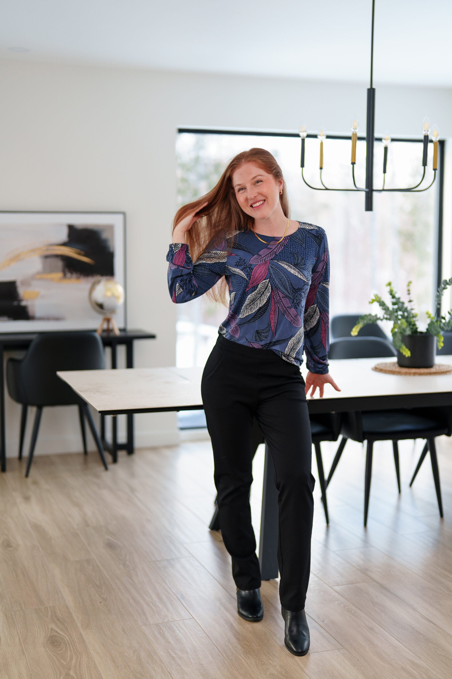 A woman wearing the St Charles Sweater by Rien ne se Perd in Foliage, a jewel toned patterned top with long sleeves and elastic cuffs. She is wearing it with the Murphy Pants in Black and 
standing in a modern kitchen. 
