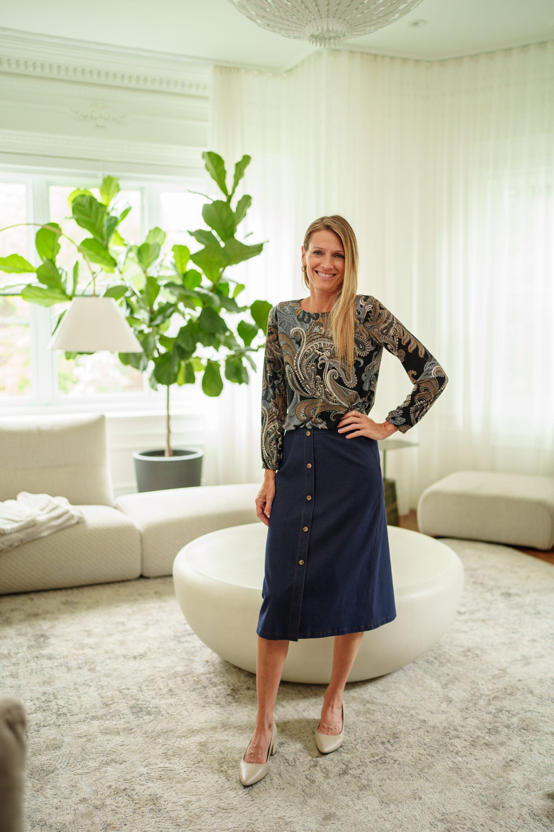A woman wearing the Nicolet Skirt from Rien ne se Perd, a blue denim midi with buttons up the front, an elasticated waist, and a centre back slit. She is wearing it with paisley top and standing in a cream coloured living room. 