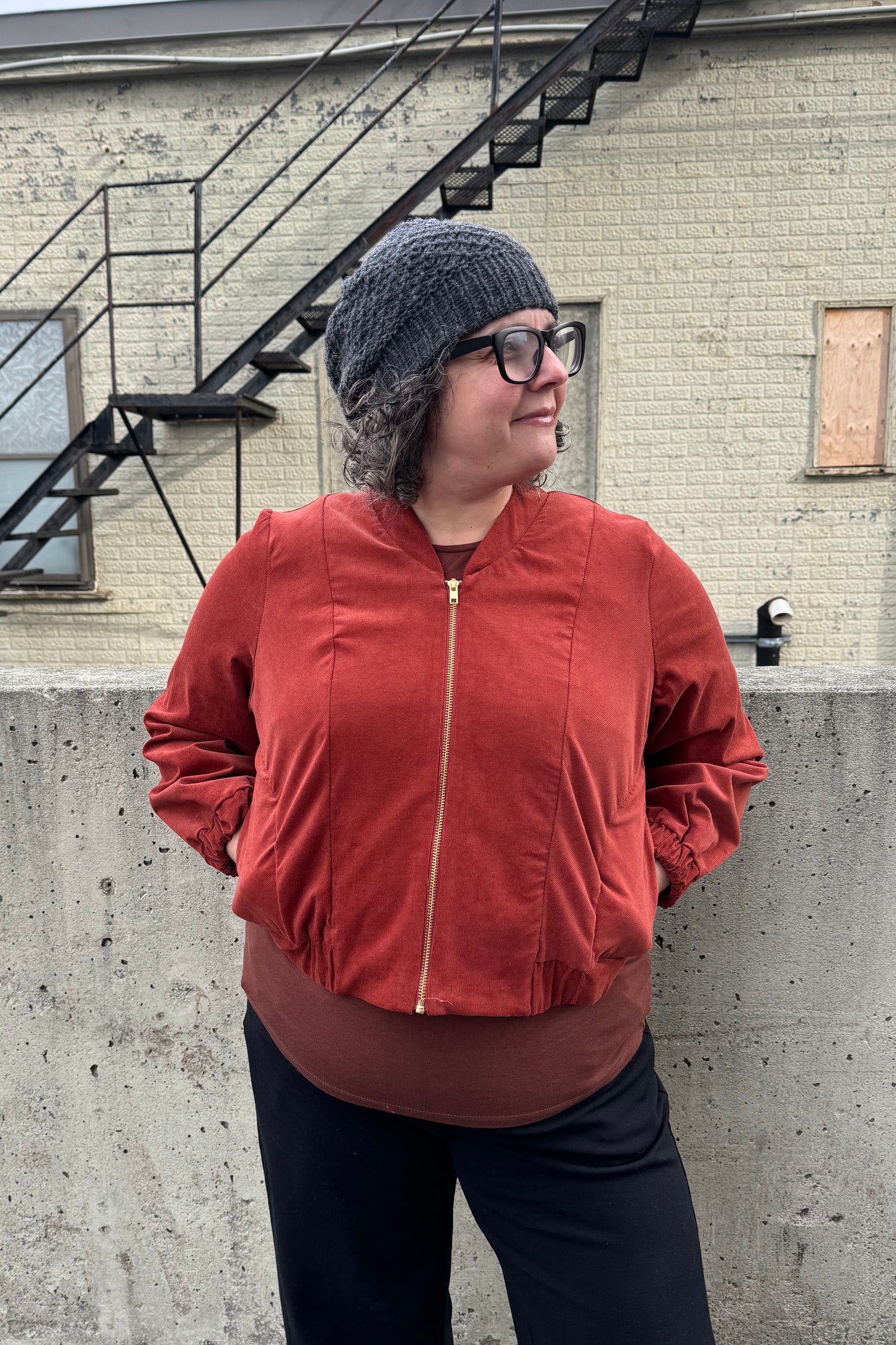 A woman wearing the Beret by Charlevoix Pure Laine in Charcoal, with the Redrawn Zip Jacket by Eve Lavoie in Terracotta Corduroy. She is standing outside. 