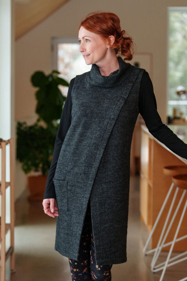 A woman wearing the long Baumier Vest by Rien ne se Perd in Charcoal, with a cowl neck, overlapping front, buttons and the shoulder and a patch pocket. She is wearing it over a black top and floral leggings and standing in a kitchen. 