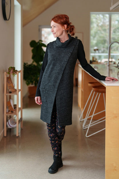 A woman wearing the long Baumier Vest by Rien ne se Perd in Charcoal, with a cowl neck, overlapping front, buttons and the shoulder and a patch pocket. She is wearing it over a black top and floral leggings and standing in a kitchen. 