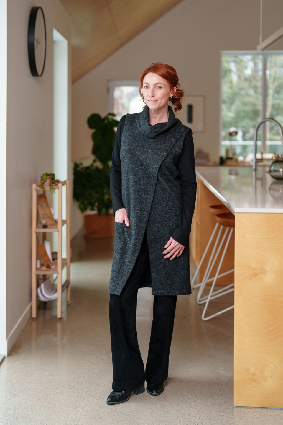 A woman wearing the long Baumier Vest by Rien ne se Perd in Charcoal, with a cowl neck, overlapping front, buttons and the shoulder and a patch pocket. She is wearing it over a black top and black pants and standing in a kitchen. 