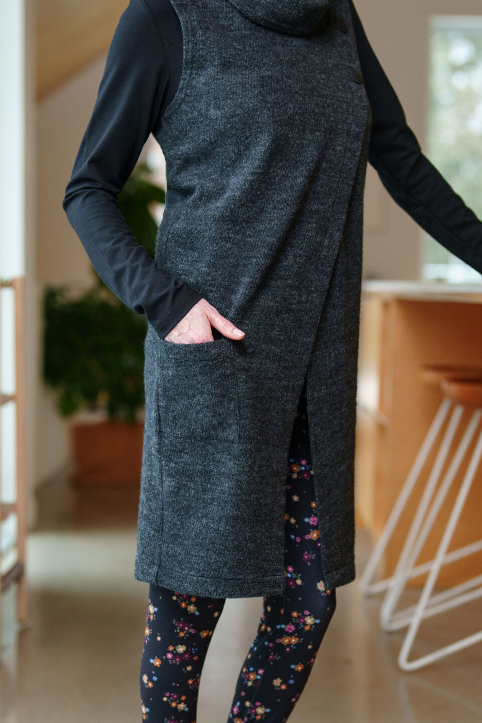 Neck down view of a woman wearing the long Baumier Vest by Rien ne se Perd in Charcoal, with a cowl neck, overlapping front, buttons and the shoulder and a patch pocket. She is wearing it over a black top and floral leggings and standing in a kitchen. 