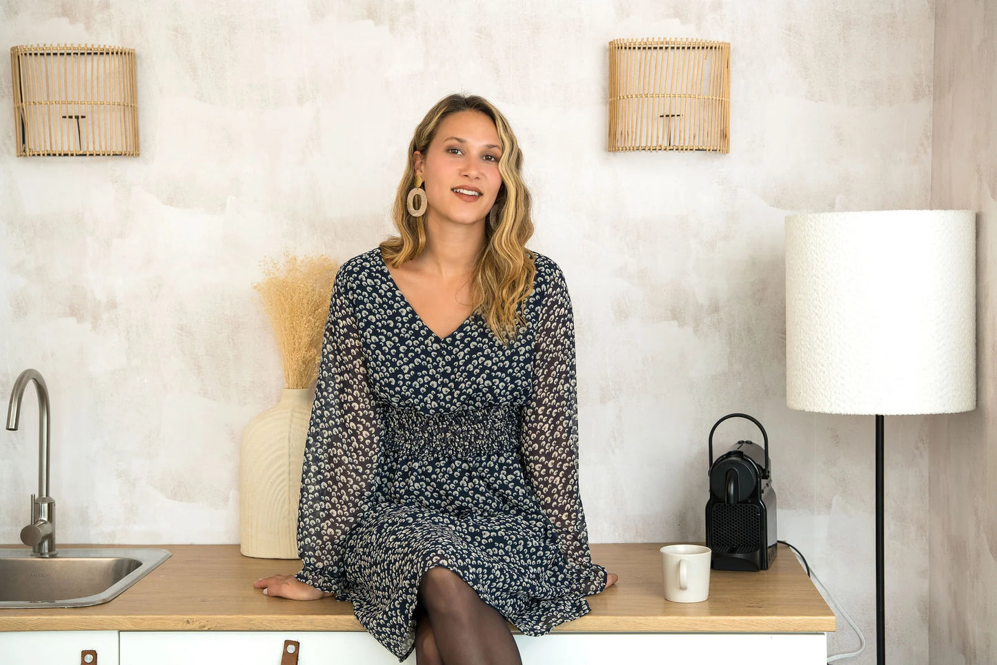A woman wearing the Spritz Dress by Cherry Bobin in Blue Pattern, featuring puff sleeves, a v-neck,  cinch waisted, and a full knee-length skirt. She is sitting on a wooden counter. 