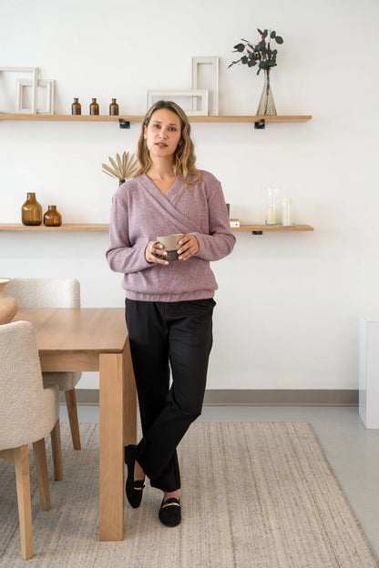 A woman wearing the Manhattan Pants by Cherry Bobin in Black, straight leg Ponte di Roma pants with a pull on waist. She is wearing them with the a mauve sweater and standing in a room with shelves. 