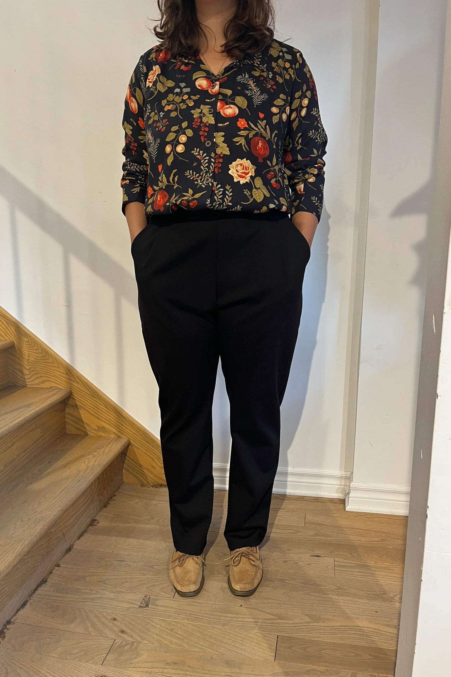 A woman wearing the Martini Blouse by Cherry Bobin in Pomegranate Apples print, a button front shirt with a classic collar. She is wearing it with black pants and standing by a staircase.
