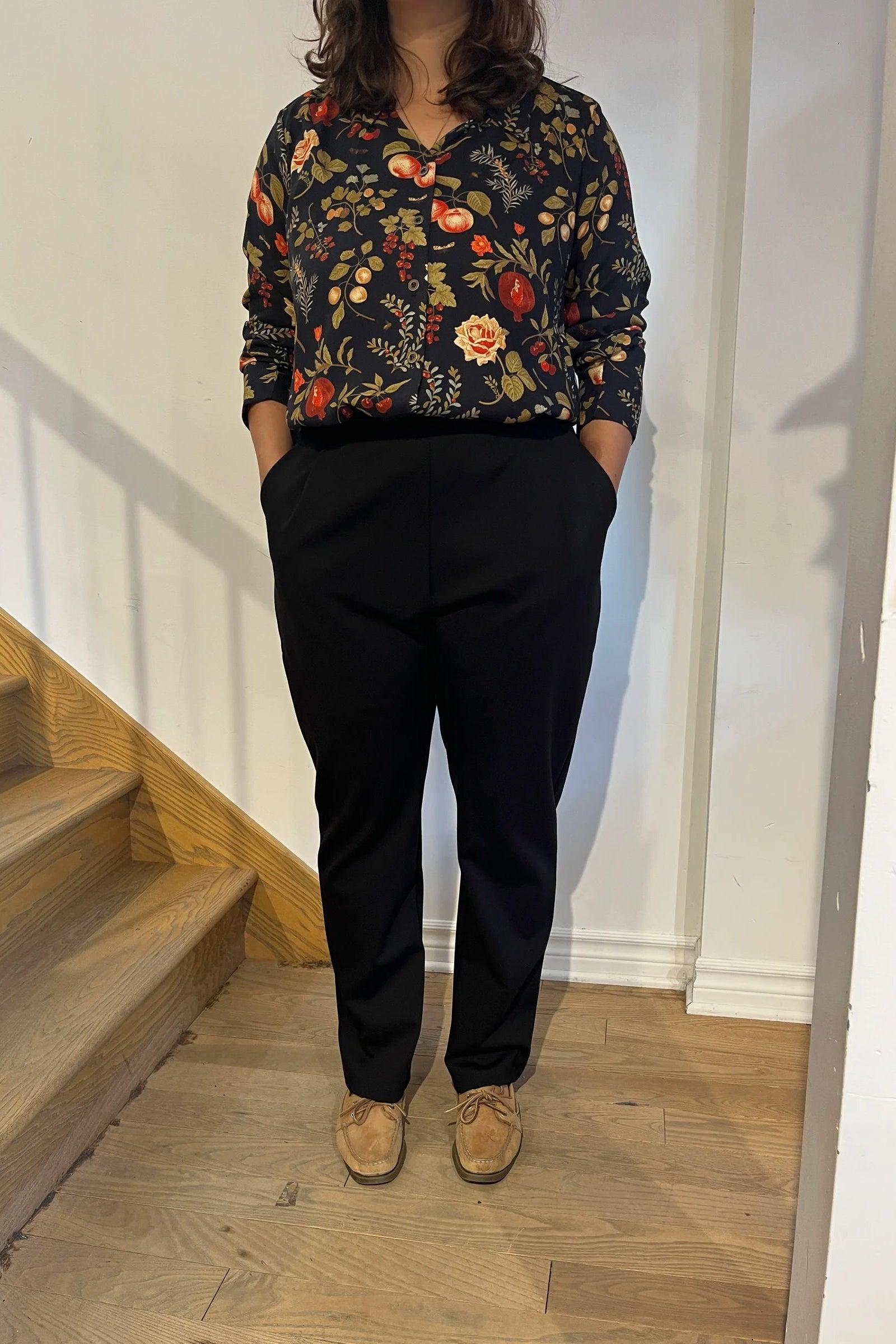 A woman wearing the Martini Blouse by Cherry Bobin in Pomegranate Apples print, a button front shirt with a classic collar. She is wearing it with black pants and standing by a staircase.

